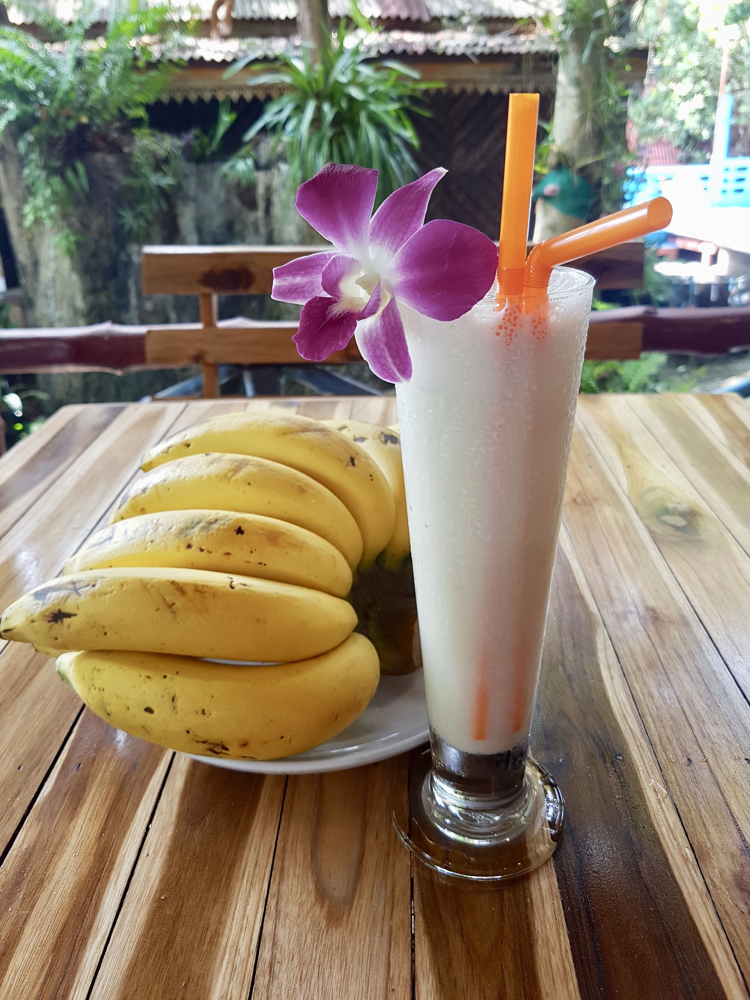 Un verre contenant une boisson blanche, orné d’une fleur violette et d’une paille orange, posé sur une table en bois.