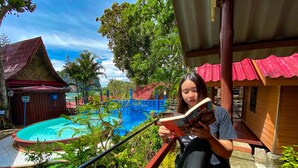 Eine Person liest ein Buch am Pool, der von einer Holzterrasse und Pflanzkübeln gesäumt ist.