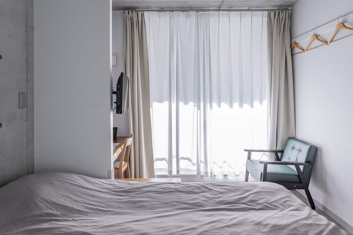 A bedroom with a bed, a chair, a TV, and a window with curtains.