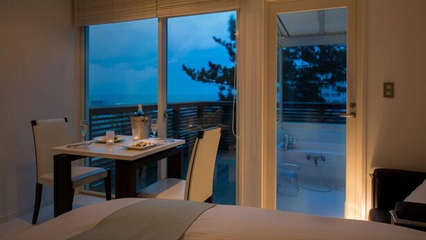 Une chambre avec un lit, une table à manger avec des chaises, un balcon donnant sur la verdure, et une porte menant à un espace extérieur.