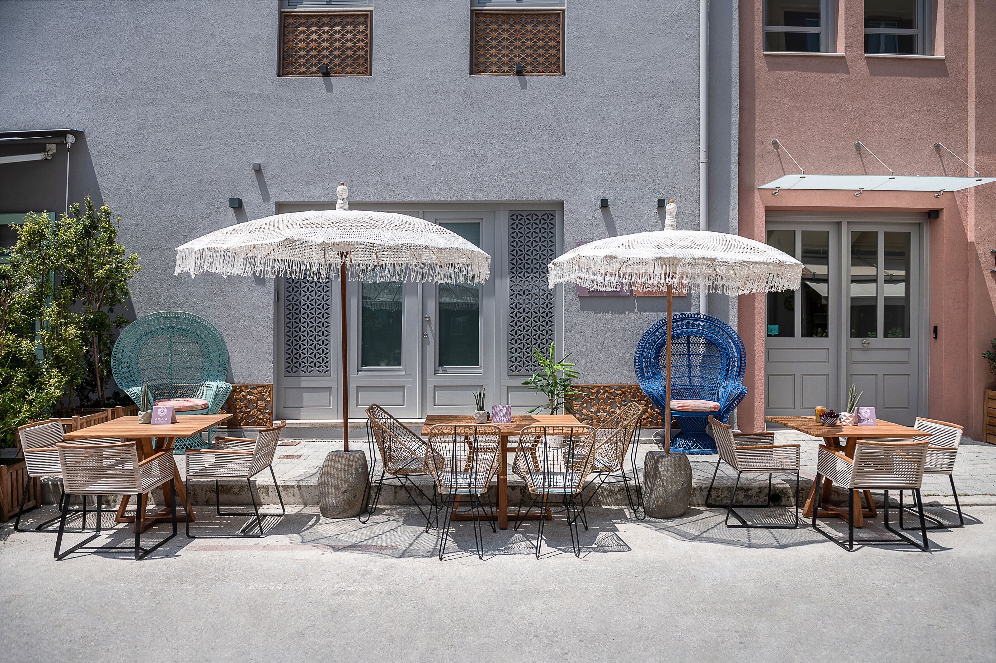Eine Terrasse im Freien mit Holztischen und Korbstühlen, weißen Sonnenschirmen und einem blauen Stuhl.