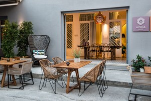 Ένας εξωτερικός χώρος με τραπέζια από ξύλο και καρέκλες από μπαμπού, ένα φυτό σε γλάστρα και μια πινακίδα που υποδεικνύει το μπουτίκ «Aisha».