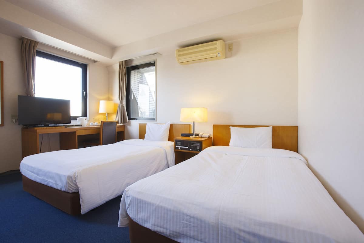 A hotel room with two beds, a desk with a television, a lamp, and an air conditioner.