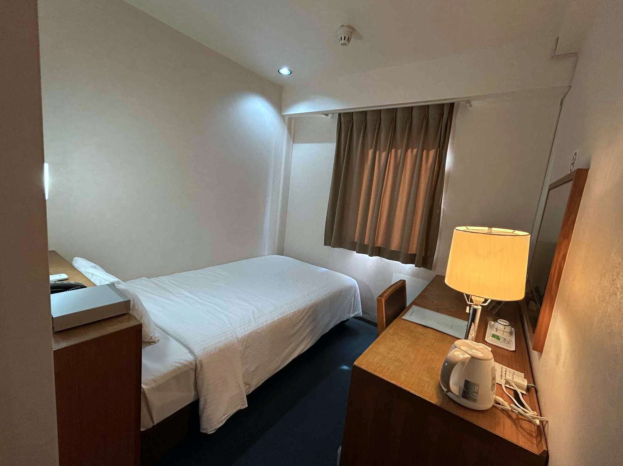 Une chambre d’hôtel avec un lit simple, un bureau avec une lampe, une chaise et une table de chevet avec une machine à café.