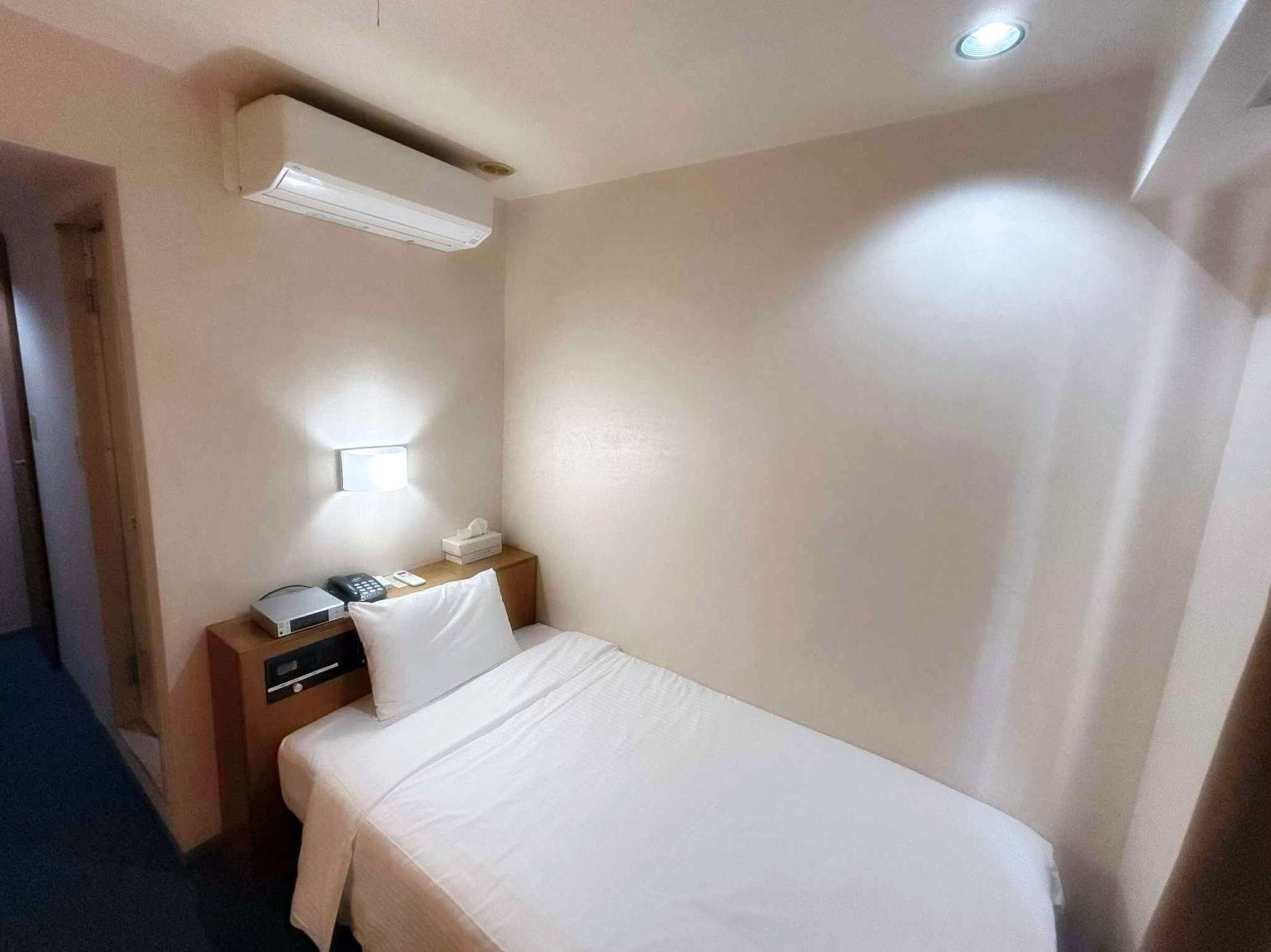 A small, neatly arranged hotel room with a single bed, a bedside table with a lamp, a telephone, and a small box, likely containing toiletries.