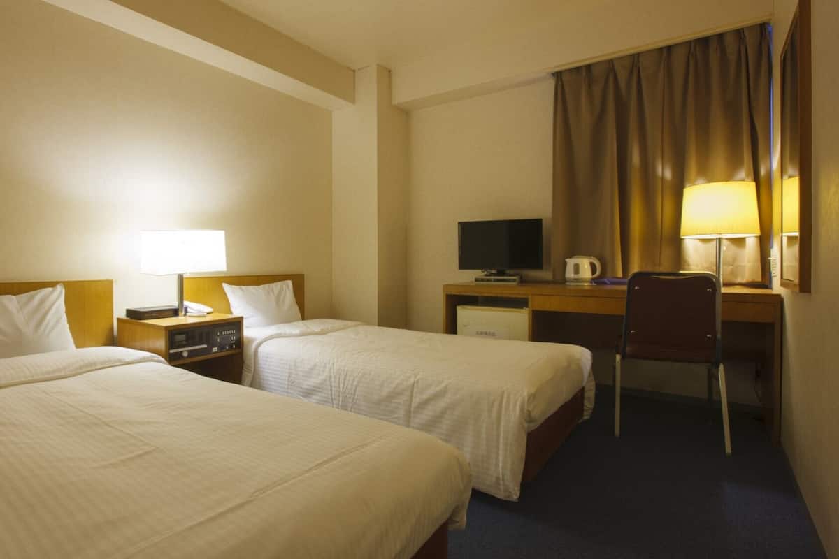 A hotel room with two beds, a desk, a chair, a television, and a lamp.