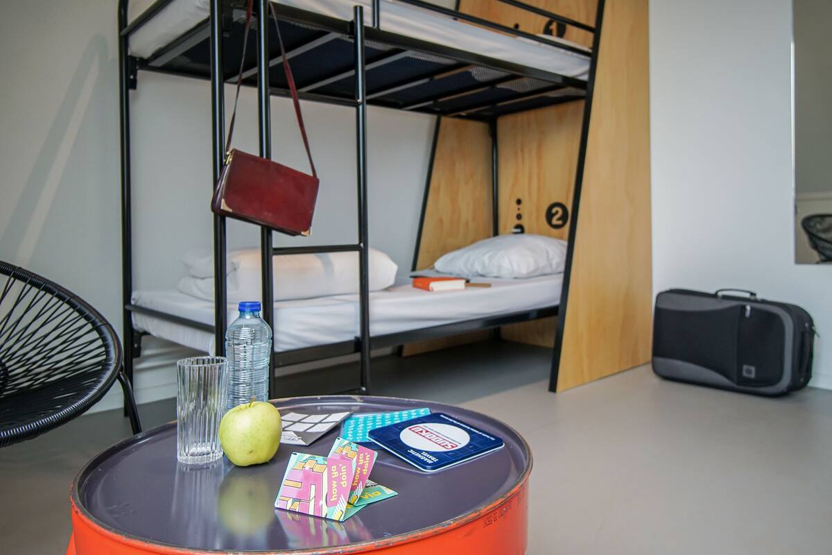 A bunk bed room with a round table, a chair, a suitcase, and a water bottle.