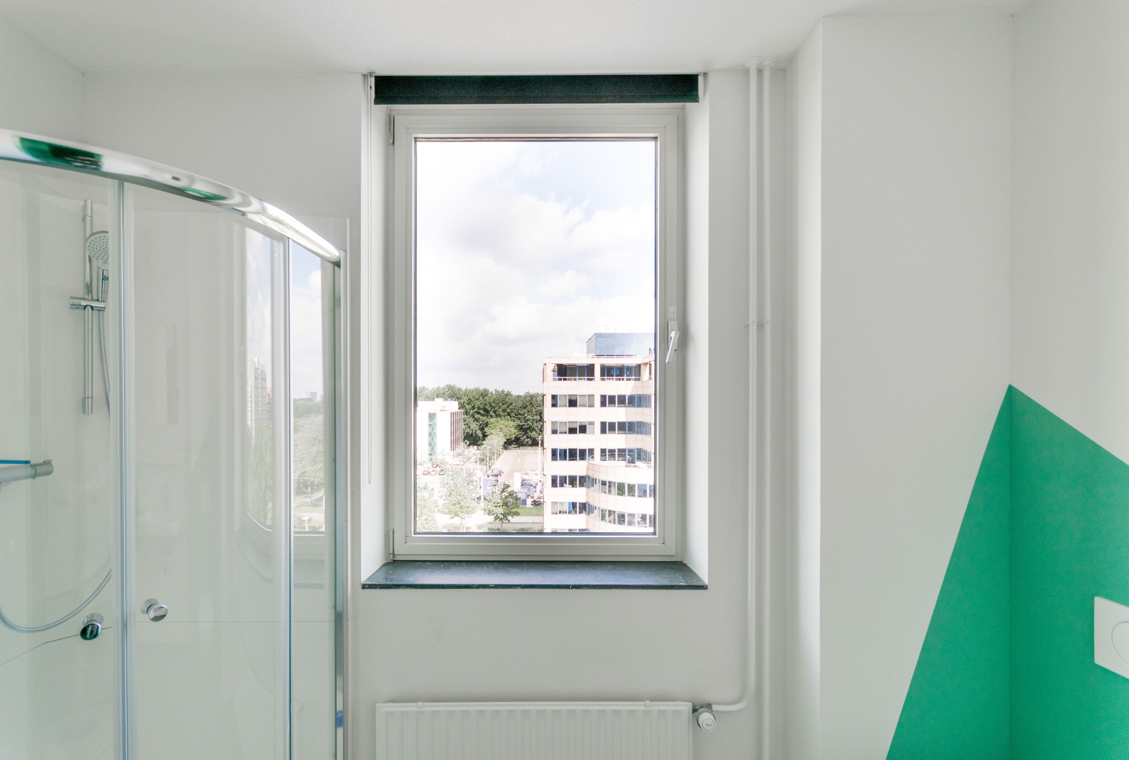 A bathroom with a glass shower enclosure, a window with a view of buildings, and a green wall.