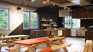 A modern kitchen with wooden tables and benches, a large window, and a view of greenery outside. - Via Amsterdam - Hostel (Diemen)