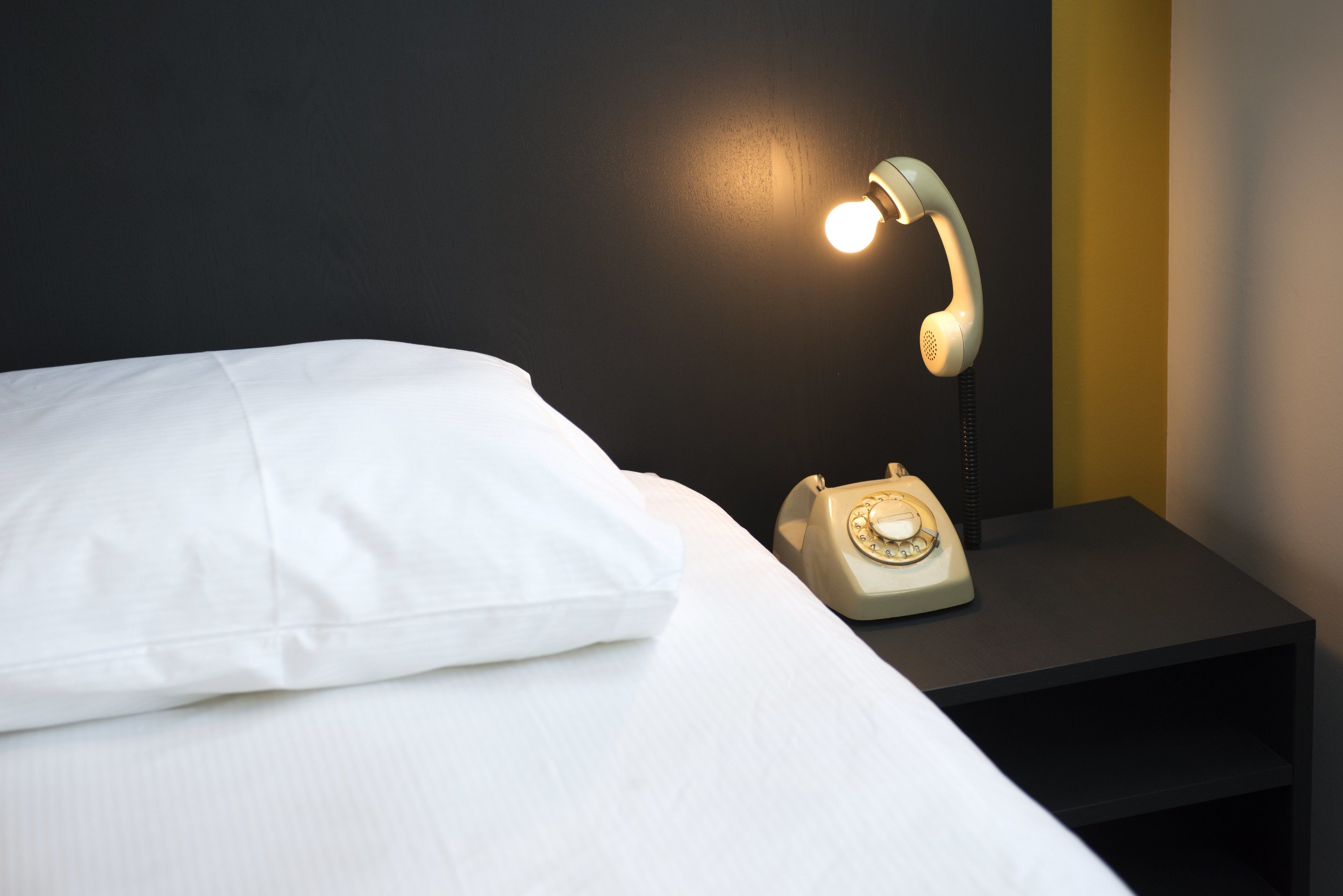 A bed with white sheets, a nightstand with a vintage-style telephone, and a wall with a dark and a yellow stripe.