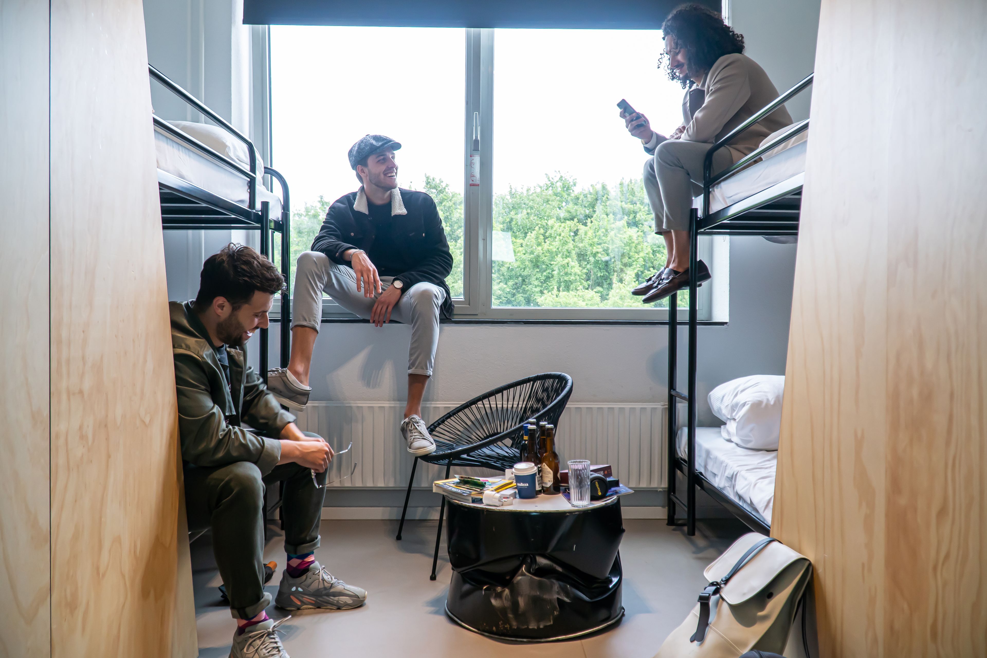 A dormitory room with bunk beds, a small table with drinks and books, and a view of trees through the window.