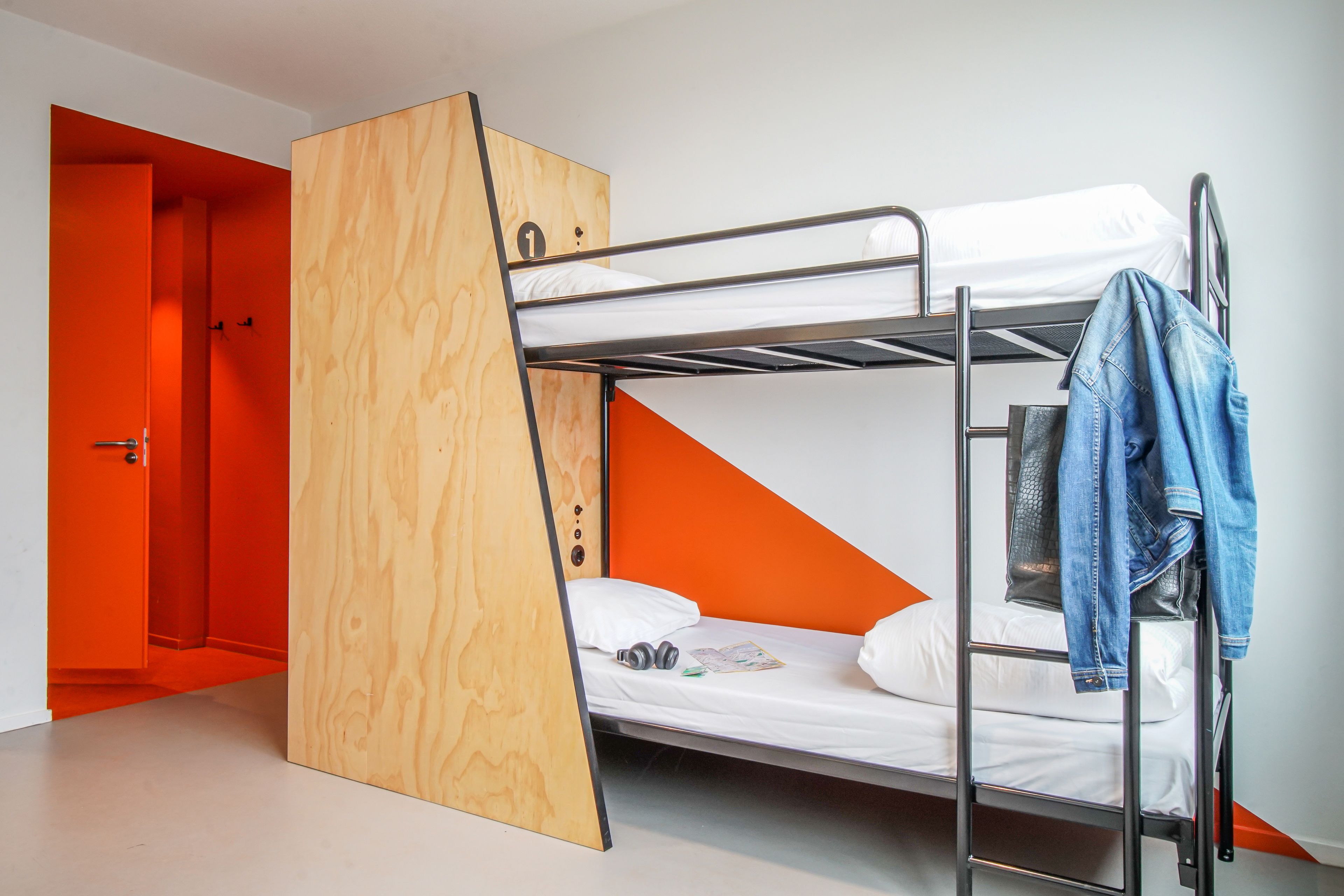 Una cama litera con escalera de madera, una cama individual con colchón y almohada blancos, y una chaqueta vaquera colgada al lado.