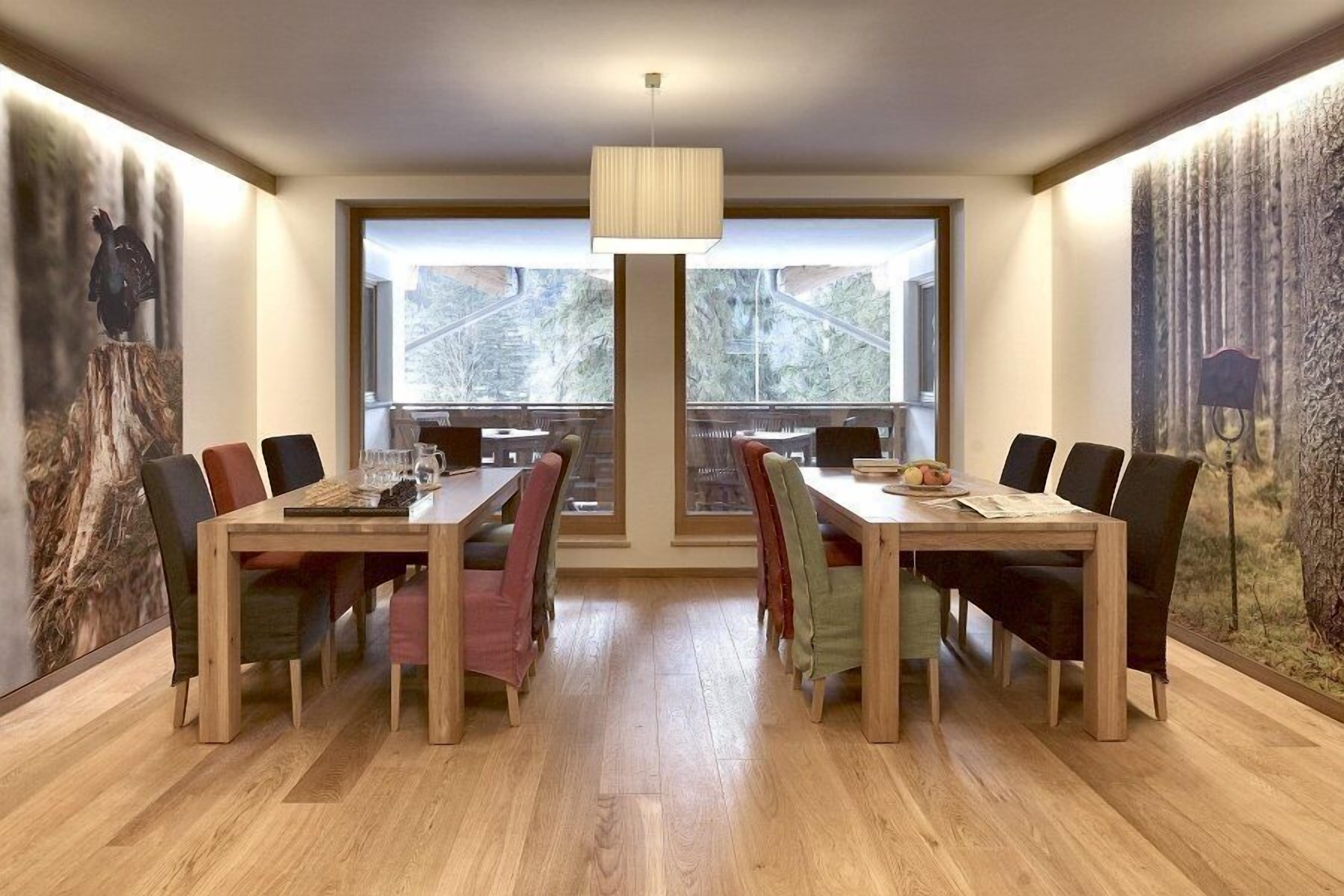 Un comedor con dos mesas de madera, sillas coloridas, una lámpara colgante y ventanas amplias con vistas a árboles.