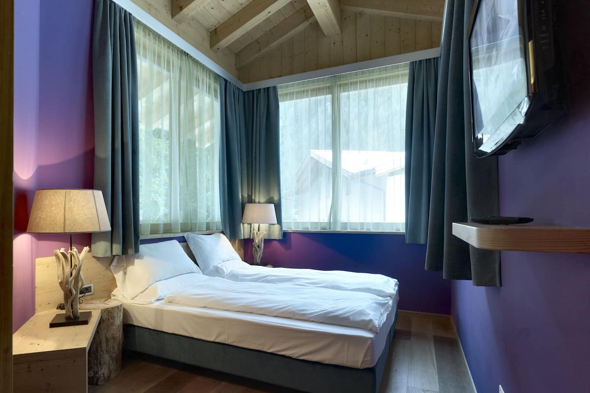 A bedroom with a bed, a nightstand, a lamp, a TV, and a window with curtains.