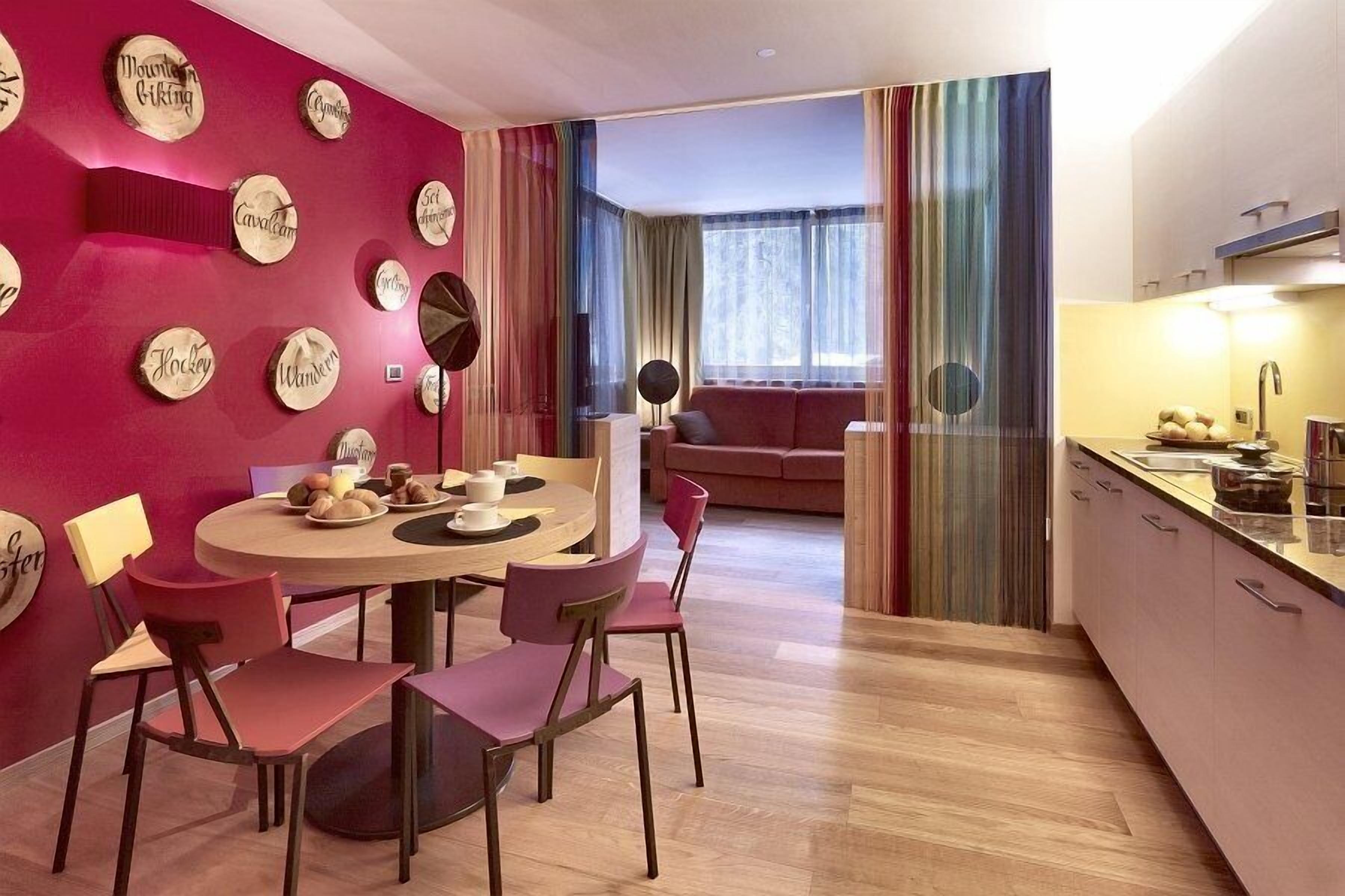 A modern living room with a round dining table, pink chairs, and a red accent wall with wooden signs.