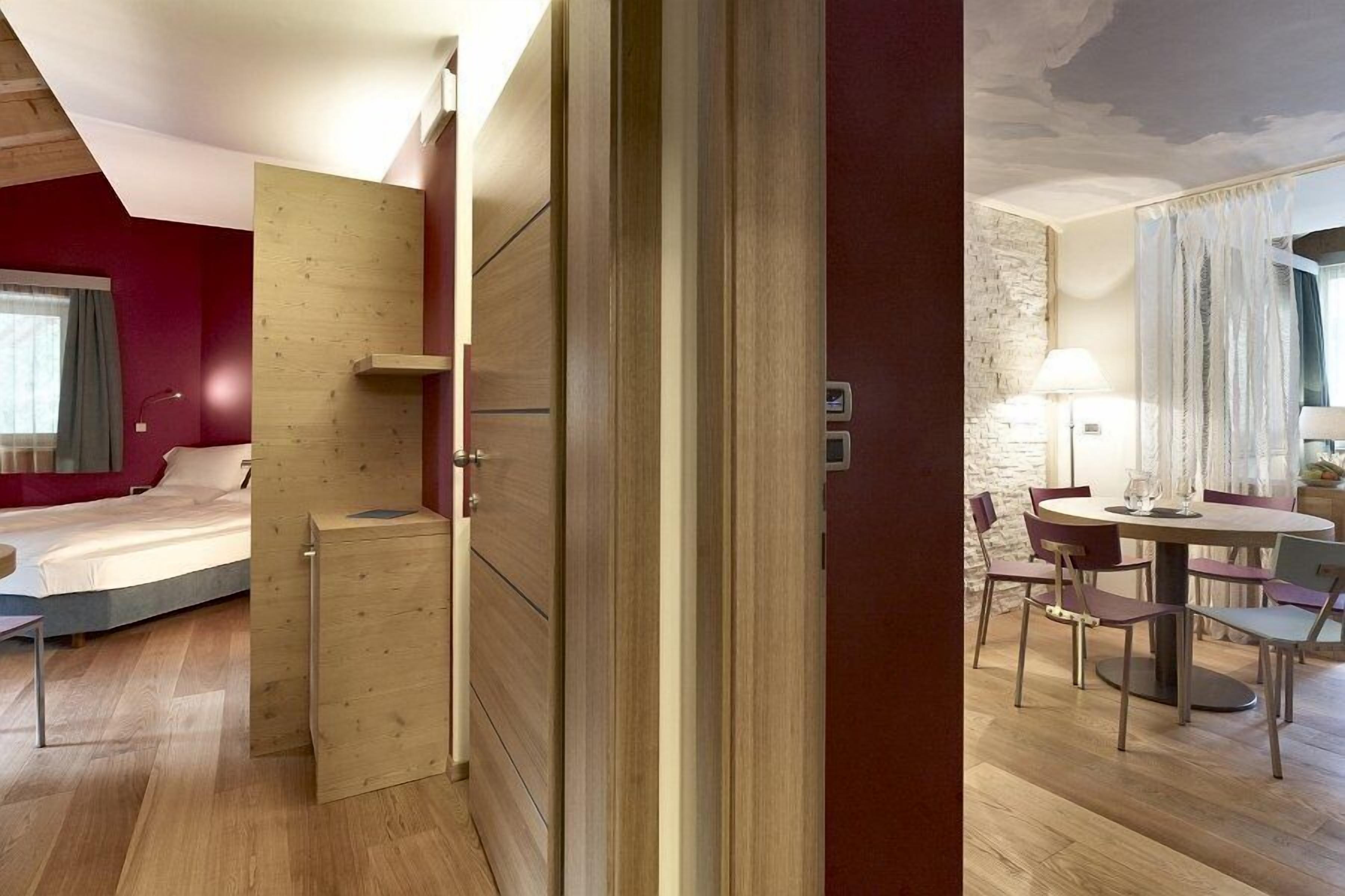 Moderni hotellihuone, jossa on sänky, puinen vaatekaappi sekä ruokailutila, jossa on tuoleja ja pöytä.