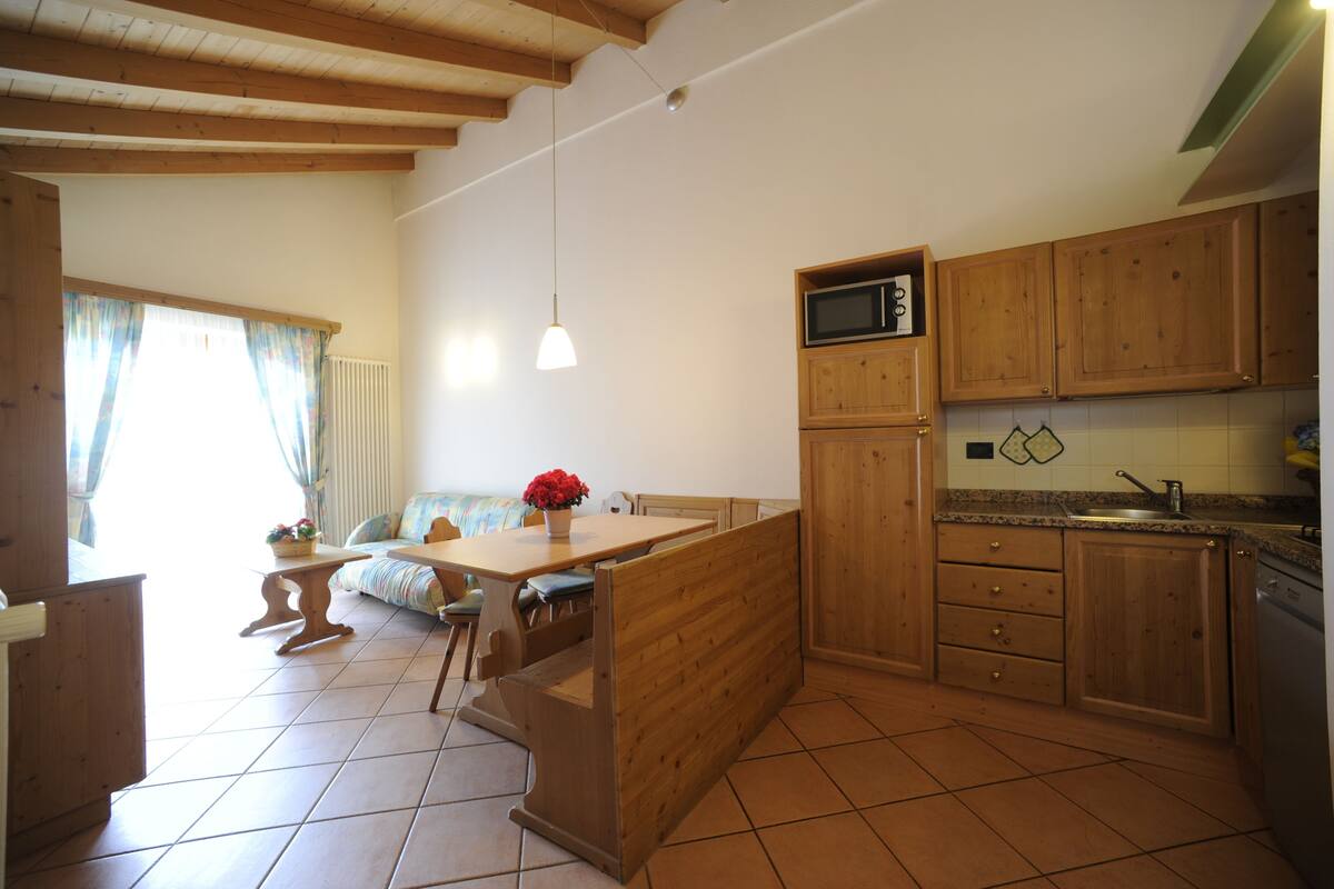 A kitchen with wooden cabinets, a microwave, and a sink. There is a dining table with chairs and a bench.