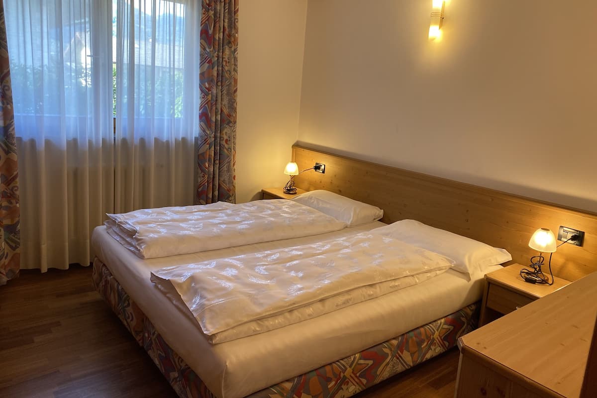 A double bed with white linens, two bedside tables with lamps, a wooden headboard, and a window with sheer curtains.