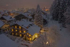 A snow-covered residential area with houses and trees, featuring a prominent house with a balcony and illuminated windows. - Residence Lagorai (Predazzo)