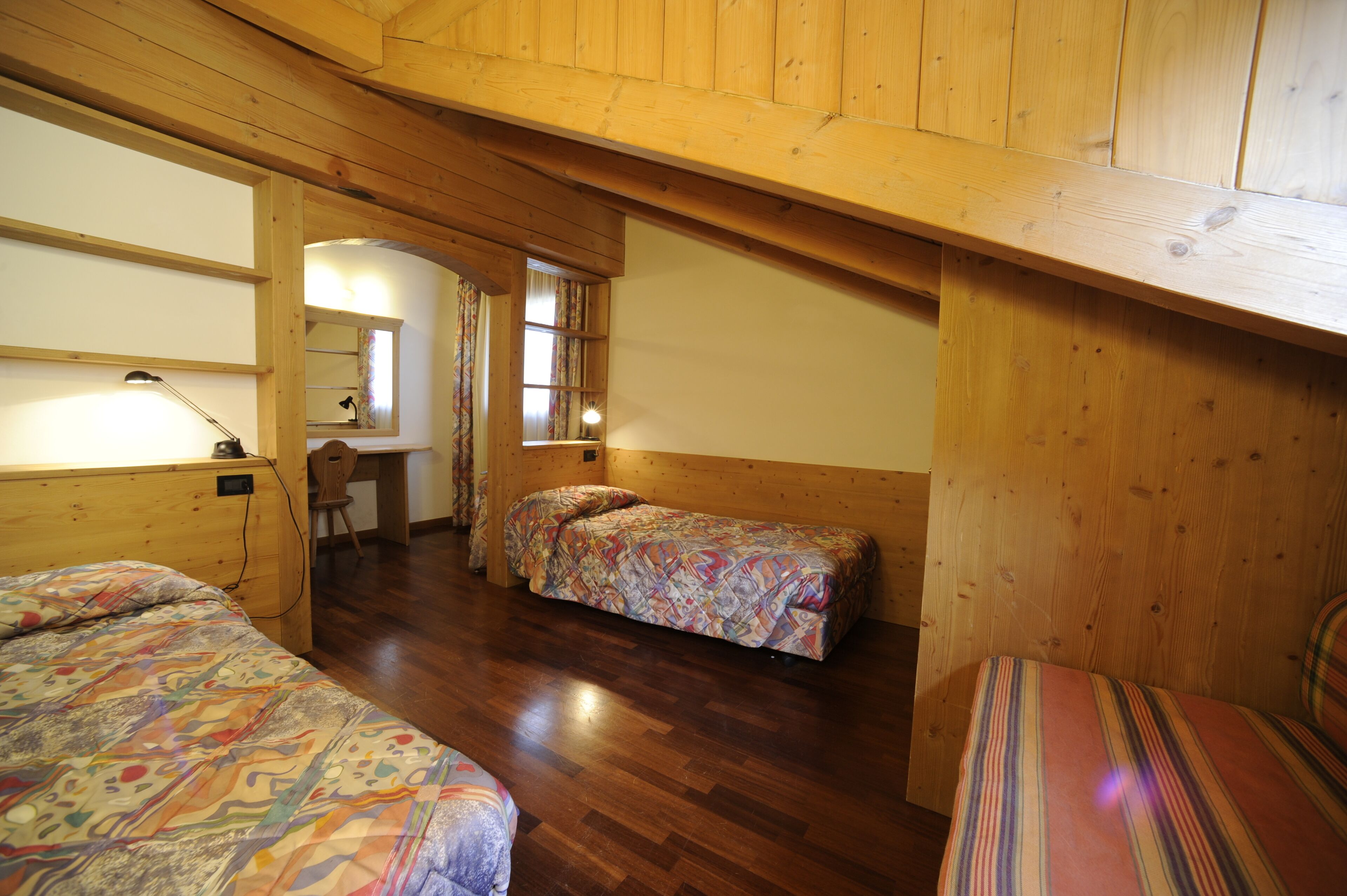 Een kamer met houten wanden en een schuin dak, met twee bedden met beddengoed met een patroon, een bureau met een stoel en een spiegel.