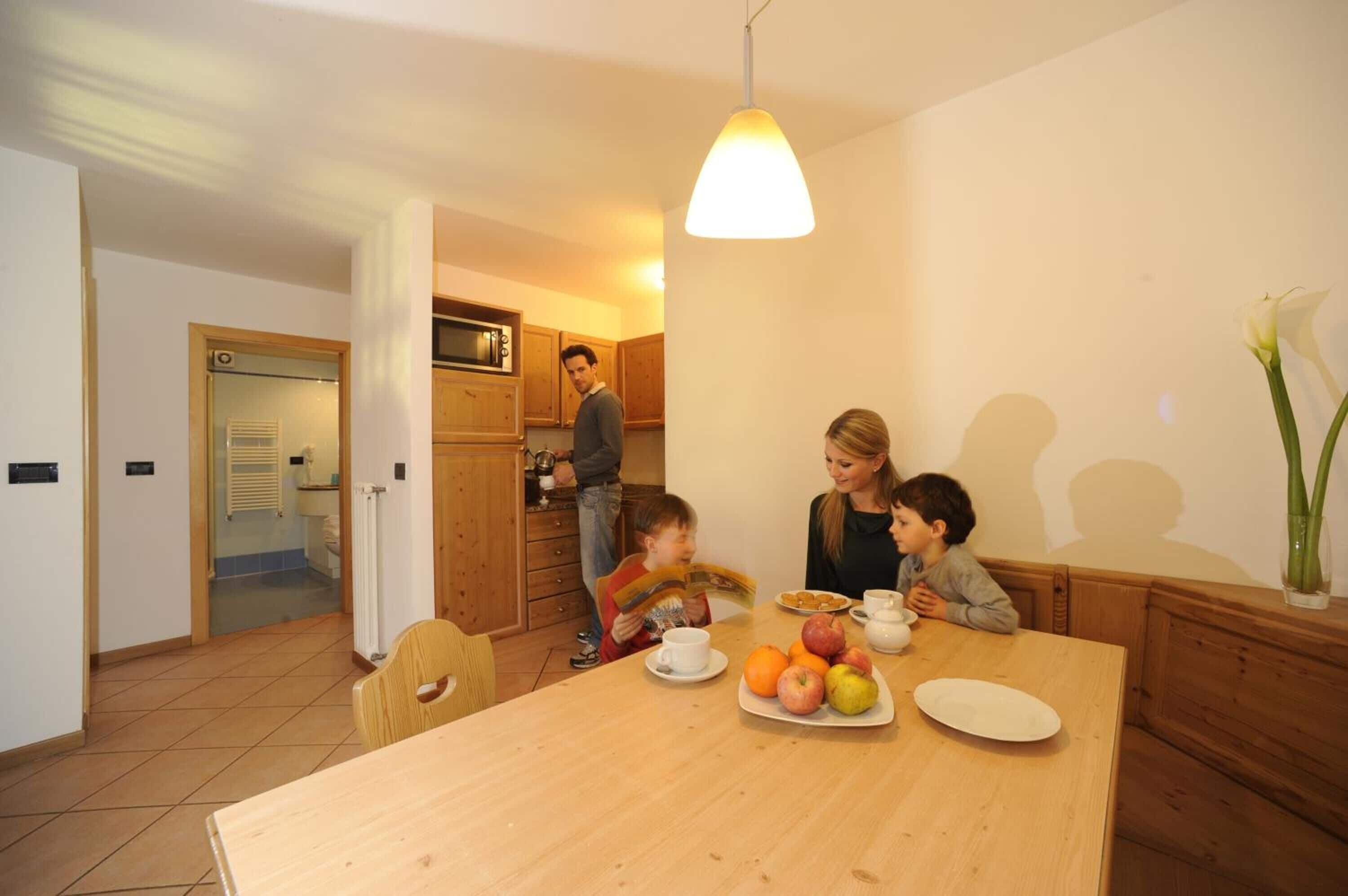 A dining area with a wooden table set for four, a kitchen in the background, and a hallway leading to a bathroom.