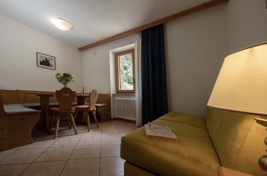 A room with a wooden table, chairs, a sofa, a window with curtains, a radiator, and a picture on the wall.