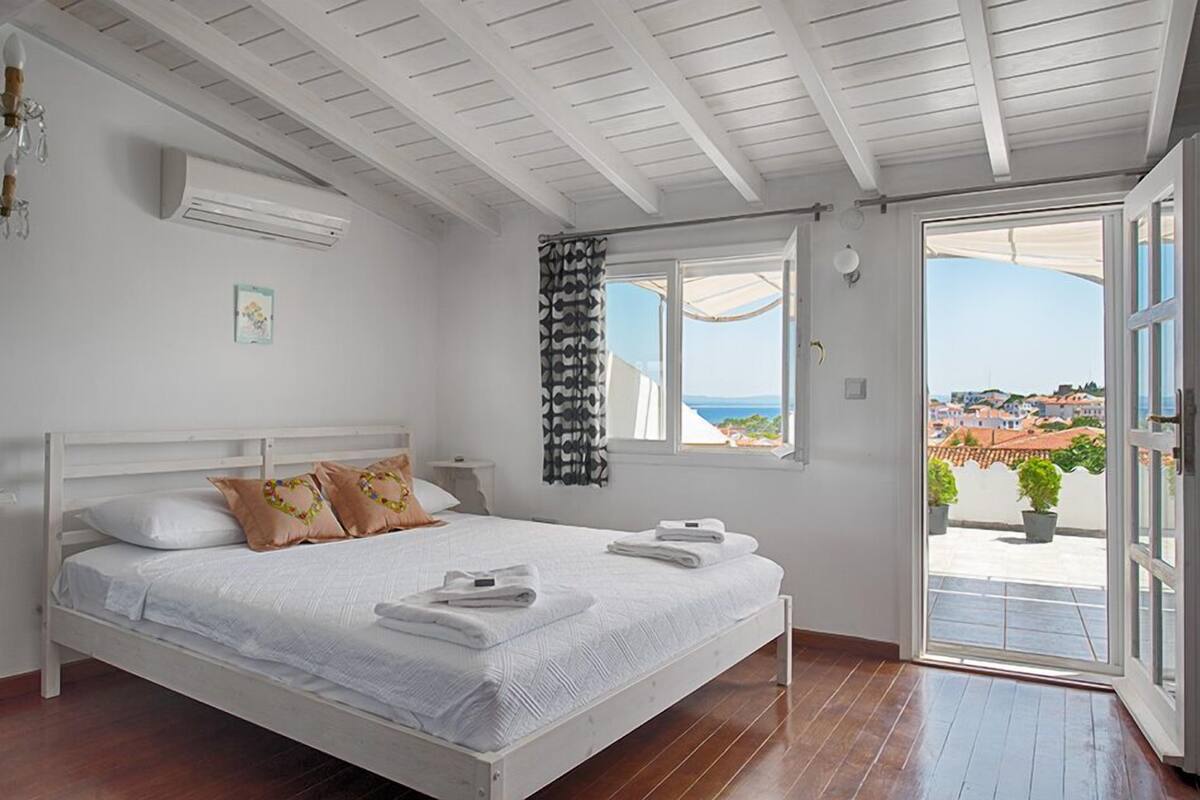 A bedroom with a white bed, a view of the sea, and a balcony with potted plants.