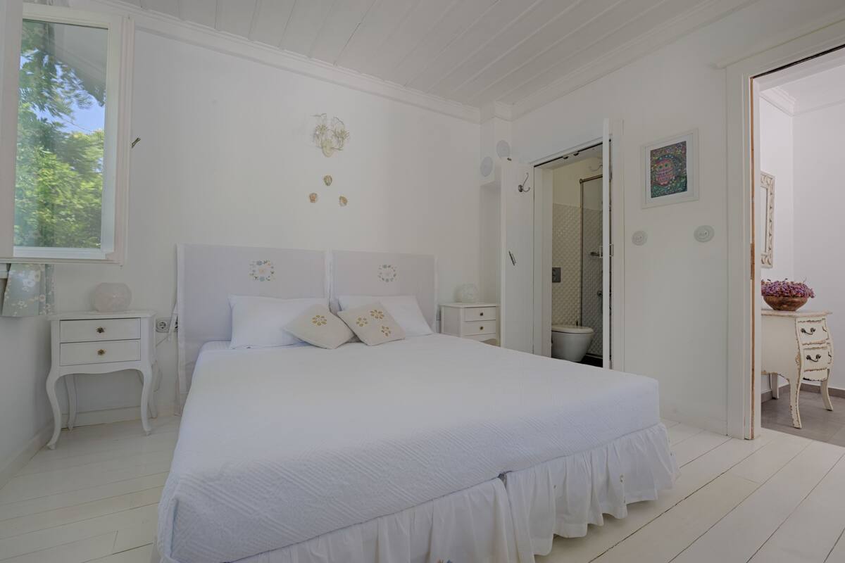 A white bedroom with a large bed, a bedside table, a nightstand, and a door leading to another room.