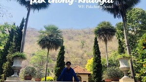 A person standing on a balcony with a view of a mountainous landscape and palm trees. - WE Pakchong Hostel (Pak Chong)