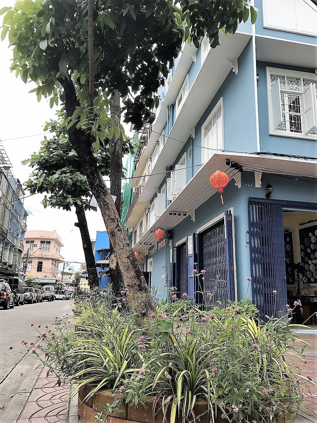 Un immeuble à plusieurs étages avec une façade bleue, un arbre devant et un parterre de fleurs roses.