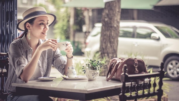 A person seated at an outdoor cafe table with a cup, a small pot, and a handbag. - The Unforgotten B&B (Bangkok)