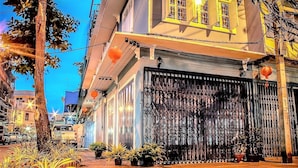 A street view with a building featuring a closed metal gate, potted plants, and a tree. - The Unforgotten B&B (Bangkok)