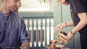 A person pouring a drink from a golden pitcher into a cup on a tray. - The Unforgotten B&B (Bangkok)