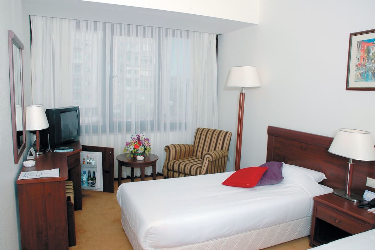 A hotel room with a bed, a chair, a desk with a television, a small table with a flower arrangement, and a lamp.