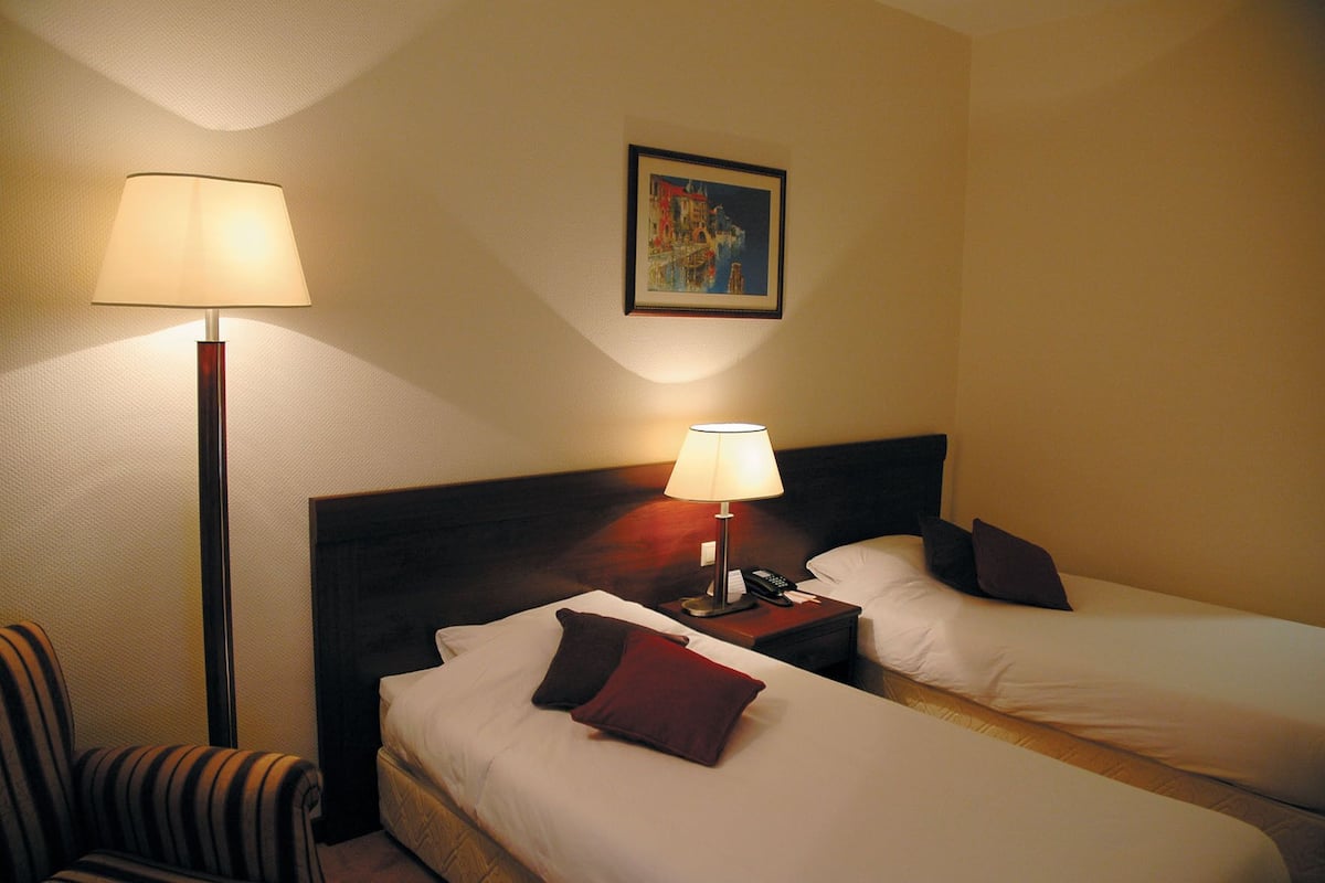 A hotel room with two beds, a nightstand with a lamp, a chair, and a framed picture on the wall.