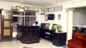 A reception area with a wooden reception desk, a man standing behind it, framed certificates on the wall, and a large window with a view of the city. - Stadium Hotel (Kumasi)