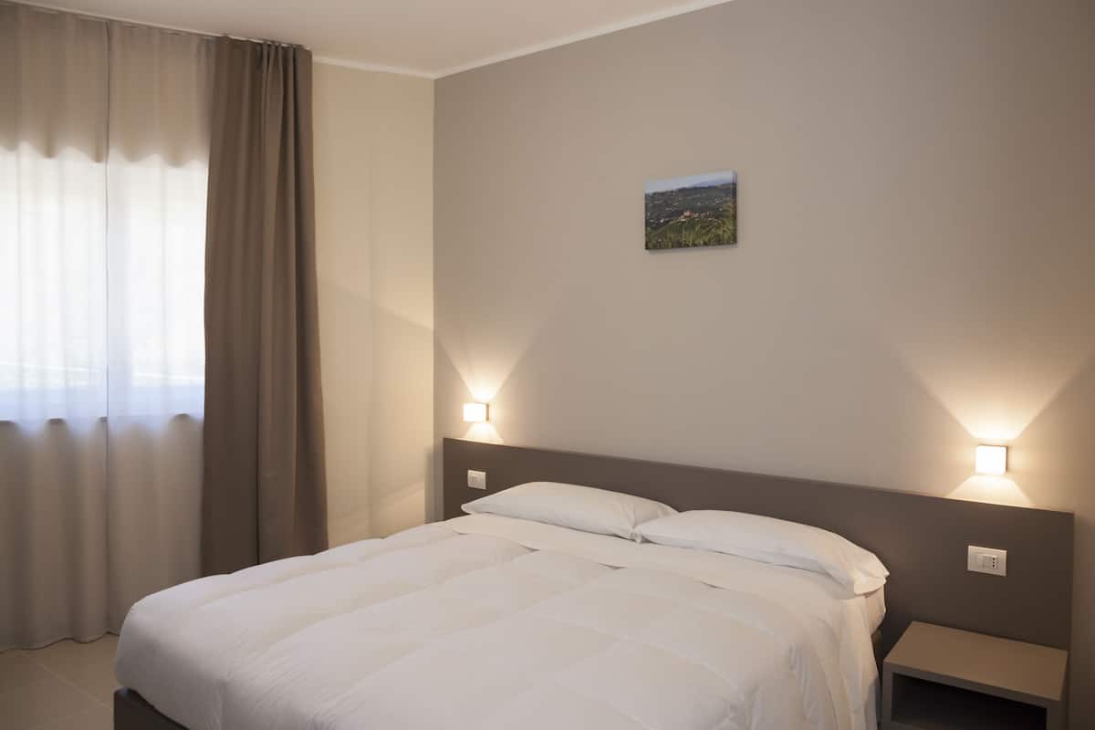Uma cama bem arrumada com roupa de cama branca, duas mesas de cabeceira com luminárias fixadas na parede e um pequeno quadro de paisagem na parede.