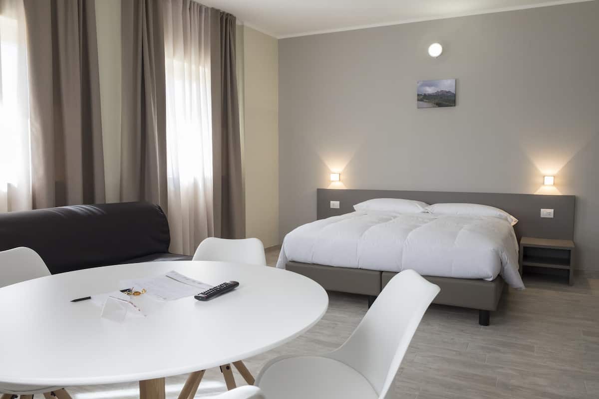 Quarto de hotel moderno com mesa de jantar redonda, cadeiras brancas, sofá cinzento e cama com roupa de cama branca, além de duas luminárias de cabeceira.
