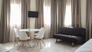 Una habitación moderna con una mesa redonda, cuatro sillas blancas, un sofá negro y un televisor de pantalla plana montado en la pared.