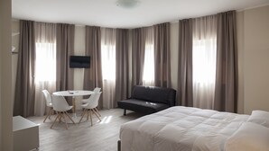 Habitación de hotel con cama, sofá, televisión, escritorio con sillas y ventanas grandes con cortinas.
