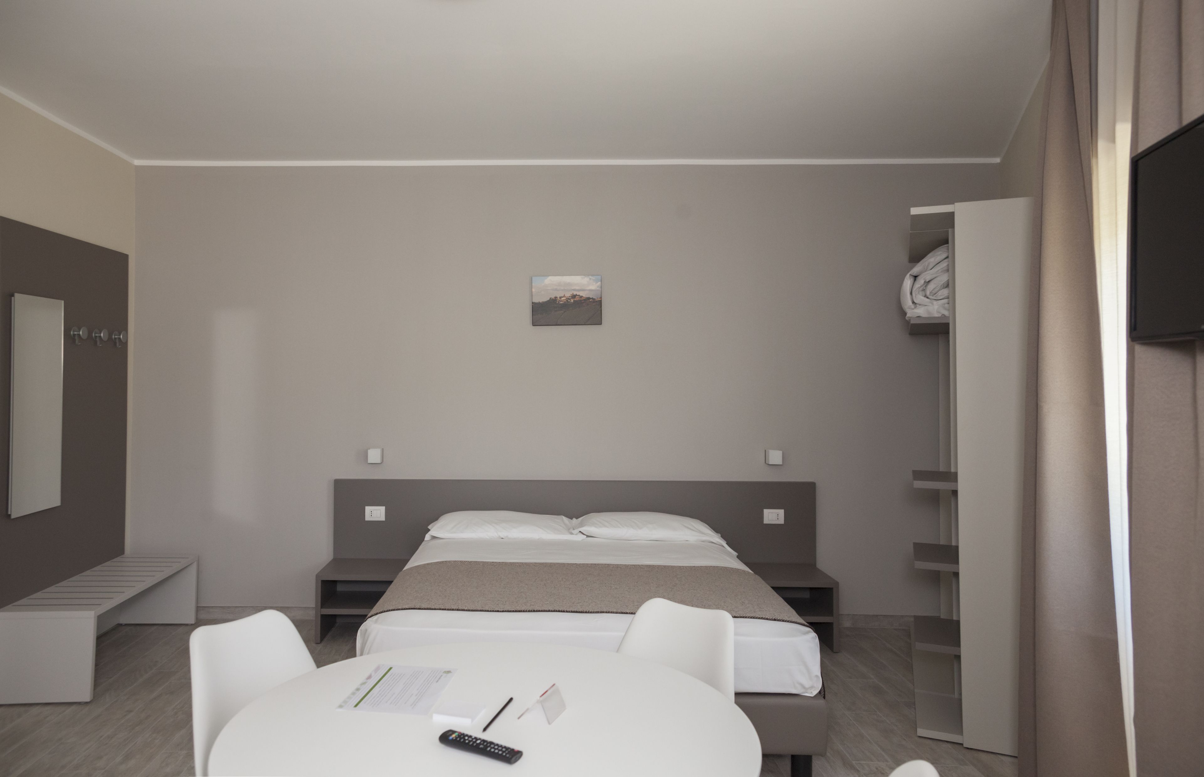 A modern hotel room with a bed, a desk, and a chair. There is a small painting on the wall and a wardrobe with a mirror.
