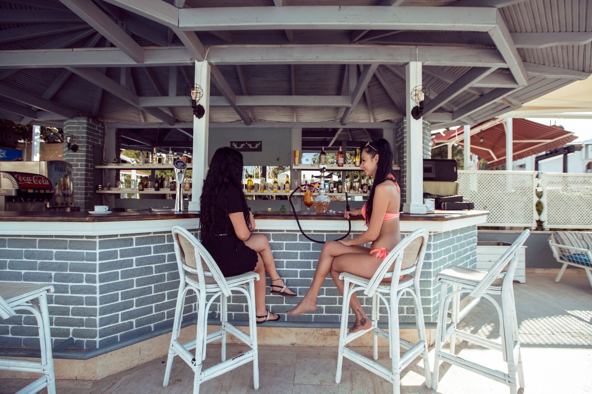 Two people seated at a bar with a brick wall backdrop, white wicker chairs, and a bar stocked with various bottles and a hookah.