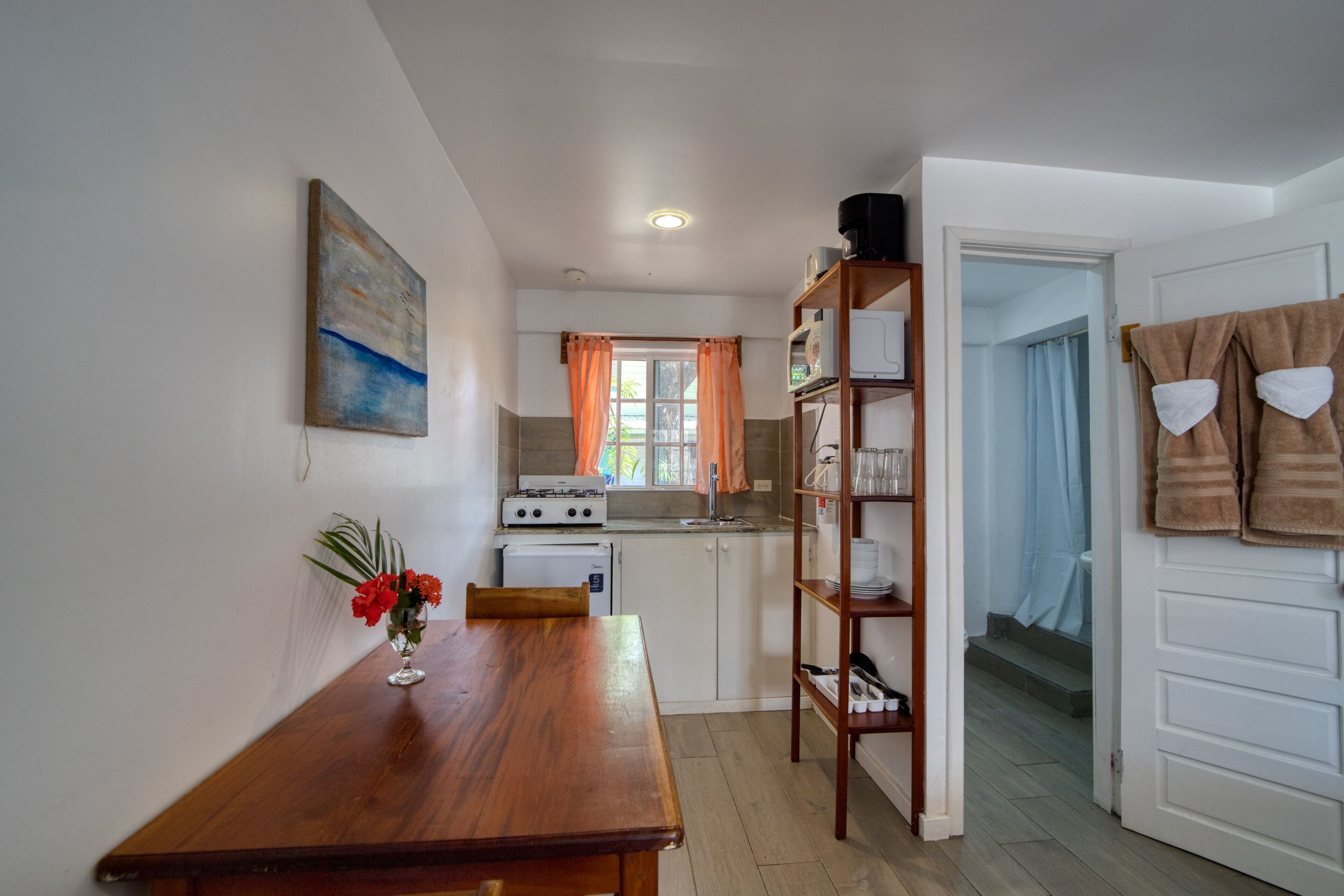 Ett kompakt kök med ett matbord, ett litet kylskåp och en spis. Det finns ett fönster med gardiner, en hylla med tallrikar och en dörr som leder till ett badrum.