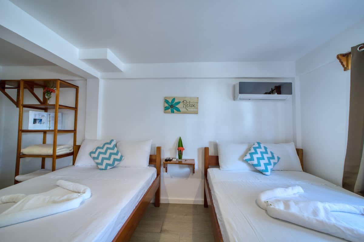 A hotel room with two single beds, a wooden shelf unit, a small bedside table with a vase and a remote control, and a wall-mounted air conditioning unit.