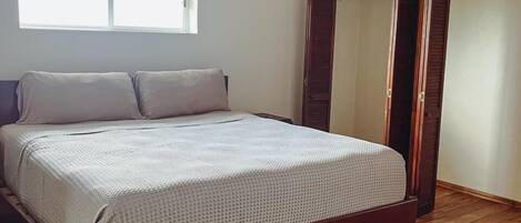 A bedroom with a wooden floor, a bed with a white bedspread, a window, and an air conditioning unit.