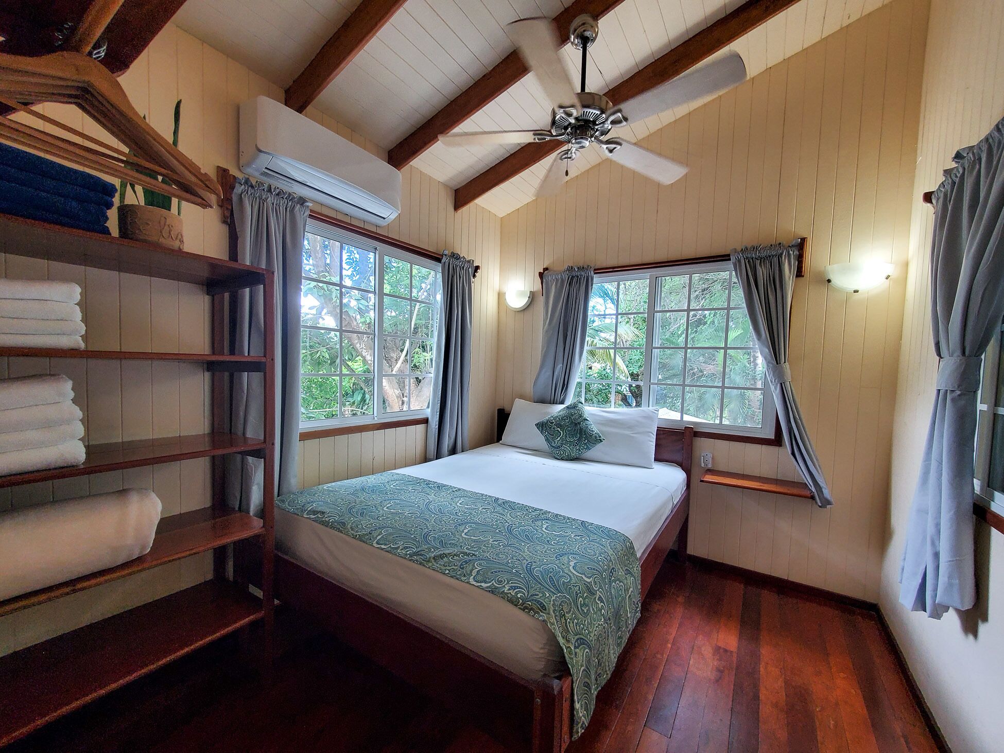 A room with a bed, wooden shelves with towels, a ceiling fan, and windows with curtains.