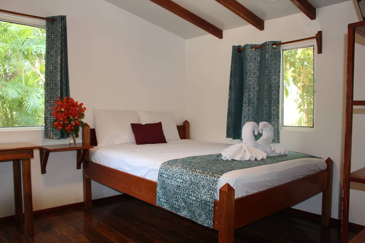 A wooden bed with a white mattress, a red pillow, and a green patterned bedspread. There is a wooden nightstand with a vase of red flowers to the left of the bed.