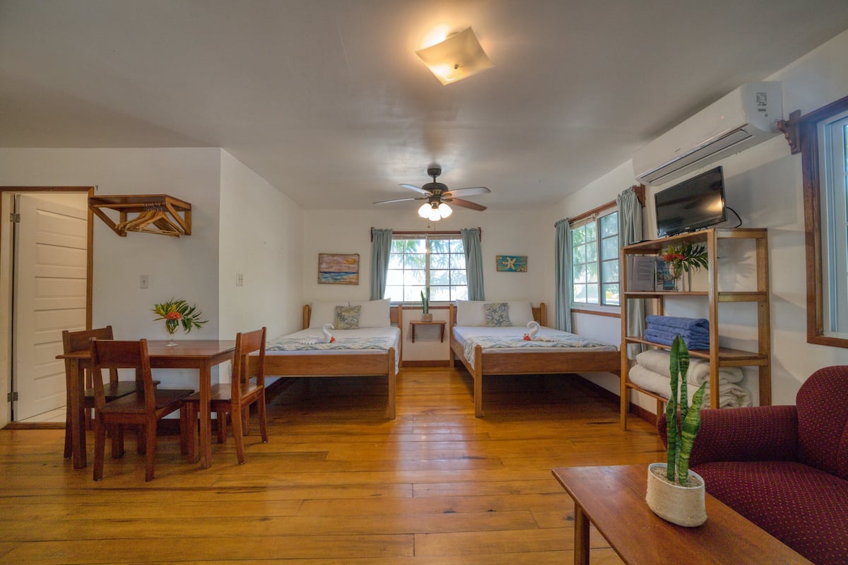 A room with two single beds, a dining table with chairs, a red sofa, a ceiling fan, and a TV on a wooden stand.