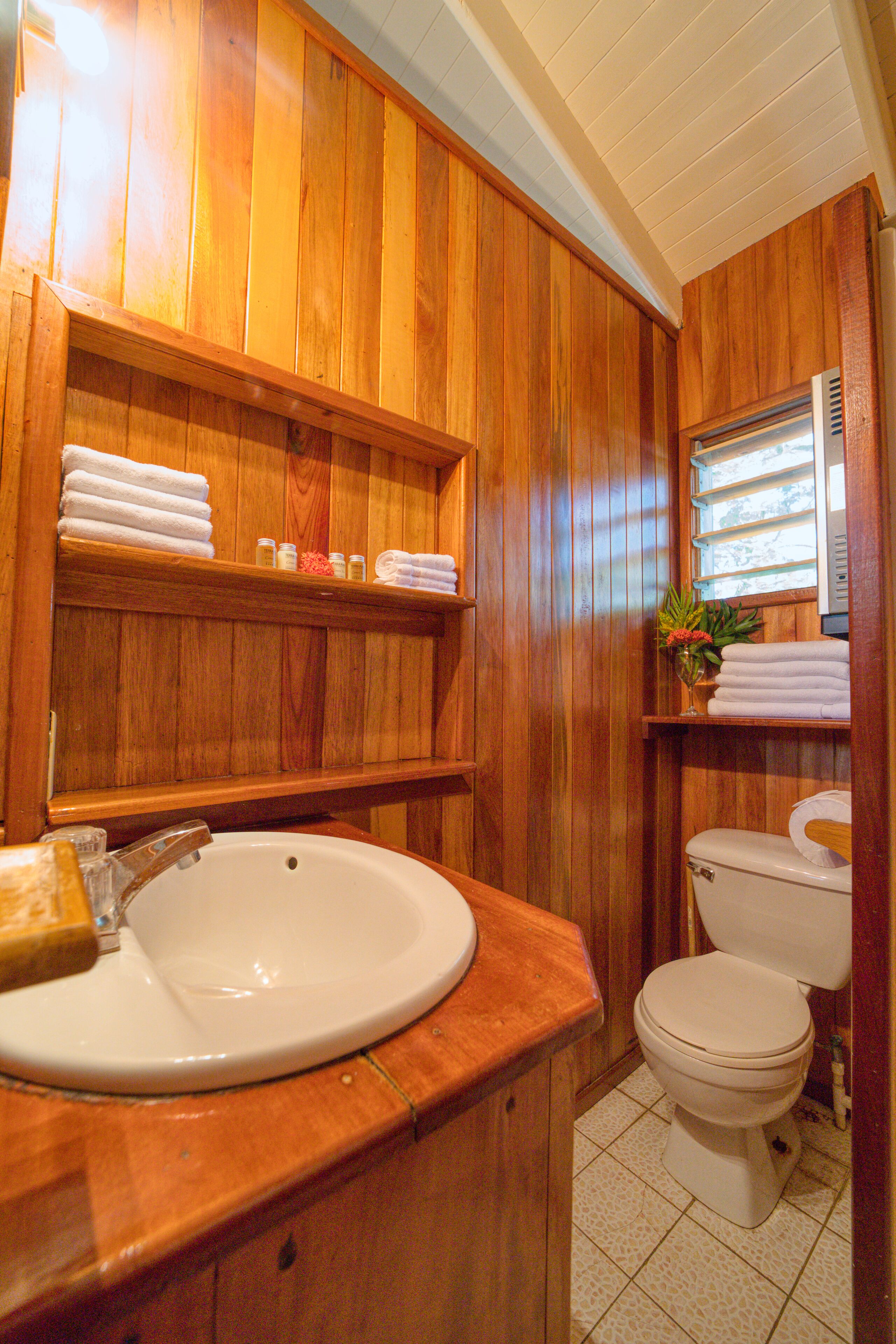 A wooden-paneled bathroom with a sink, toilet, and shelves holding towels and toiletries.