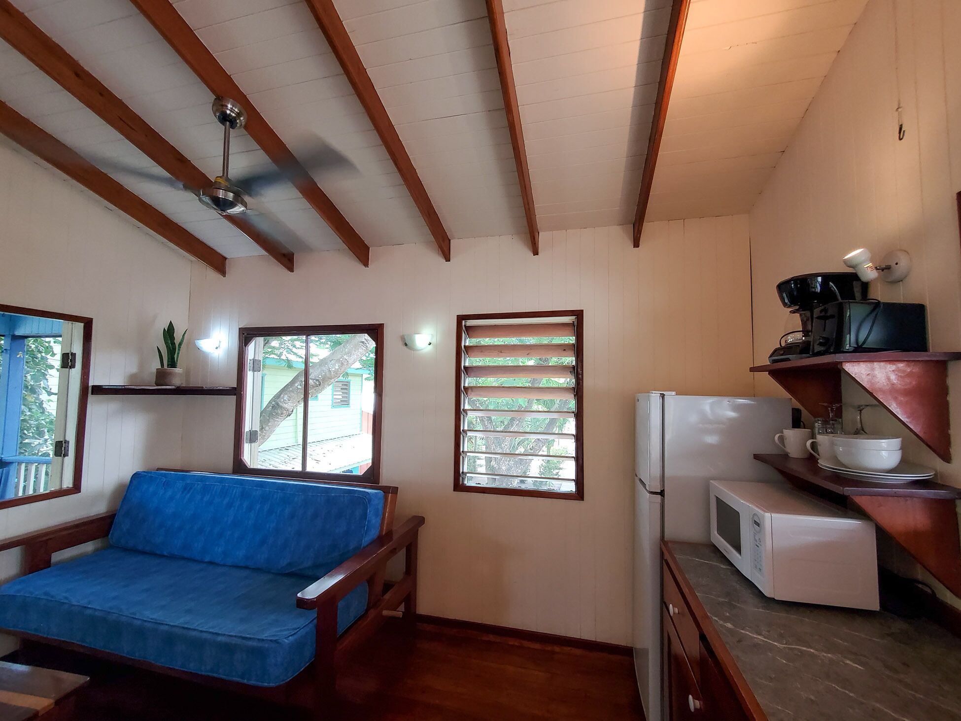 A small, cozy room with a blue sofa, a ceiling fan, a refrigerator, a microwave, and a window with blinds.
